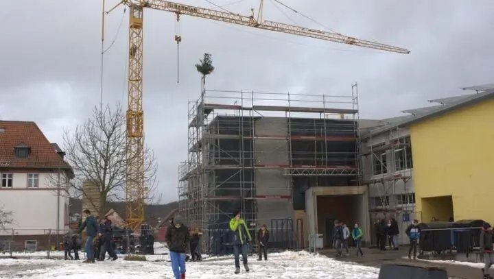 Eingerüsteter Rohbau mit Richtbaum auf dem Dach und Baukran, davor Schülerinnen und Schüler im Schnee