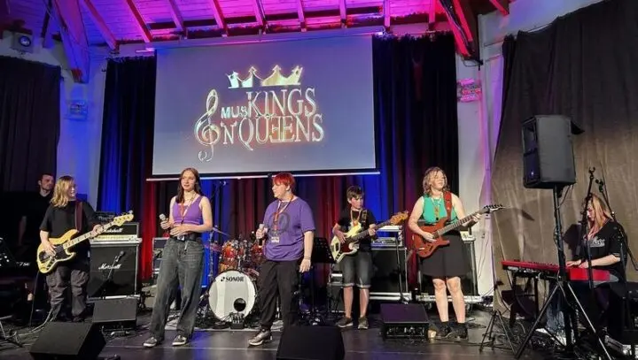 Schulband mit Gesang, Gitarren, Schlagzeug und Keyboard auf der Bühne, Leinwand mit Schriftzug „MusiKINGS'N'QUEENS“