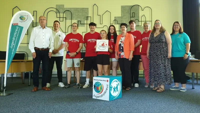 Gruppe in roten GemS-Freisen-Shirts mit Erwachsenen, eine Schülerin hält die Plakette, davor ein Jugendrotkreuz-Würfel