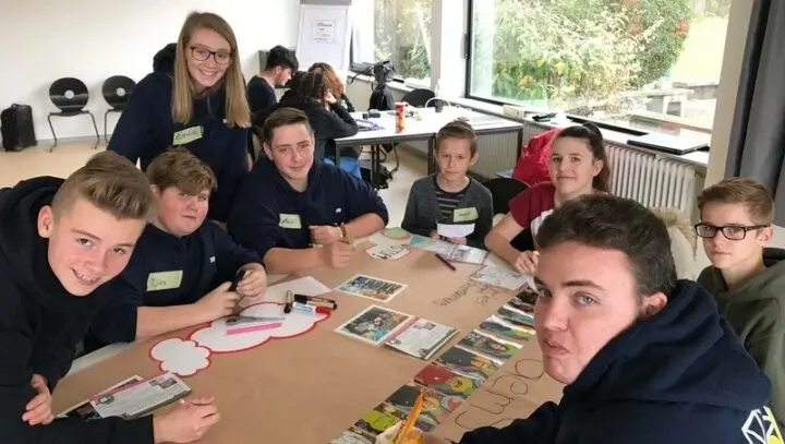 Schülervertretung mit Namensschildern arbeitet an einem Tisch mit Plakat, Bildern und Stiften
