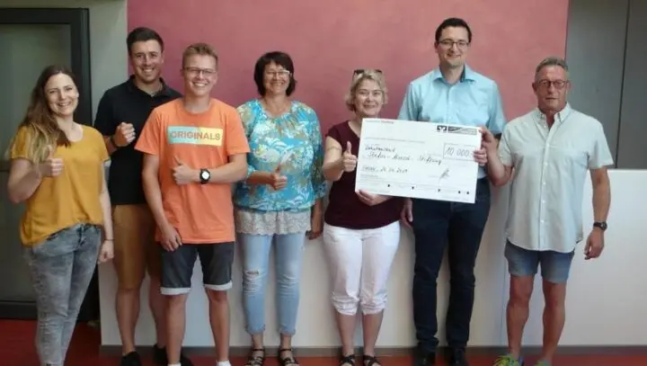 Sieben Erwachsene mit erhobenen Daumen halten einen Spendenscheck über 10.000 Euro