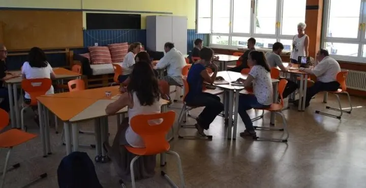 Lehrkräfte sitzen in Gruppen an Tischen in einem hellen Klassenraum mit orangefarbenen Stühlen