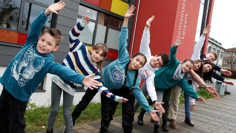 Kinder breiten vor dem Schulgebäude die Arme aus und lächeln in die Kamera