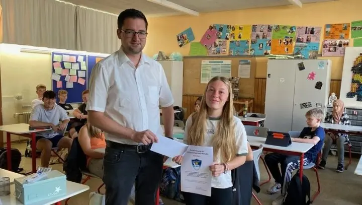 Schülerin mit Urkunde und ein Erwachsener mit Umschlag im Klassenraum