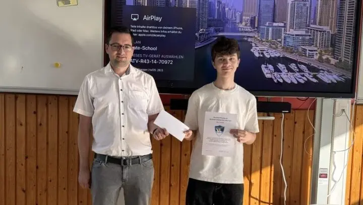 Ein Mann überreicht einem Schüler mit Urkunde einen Umschlag vor einem Bildschirm