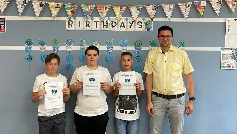 Drei Schüler halten Urkunden vor einer Klassenraumwand, daneben ein Erwachsener