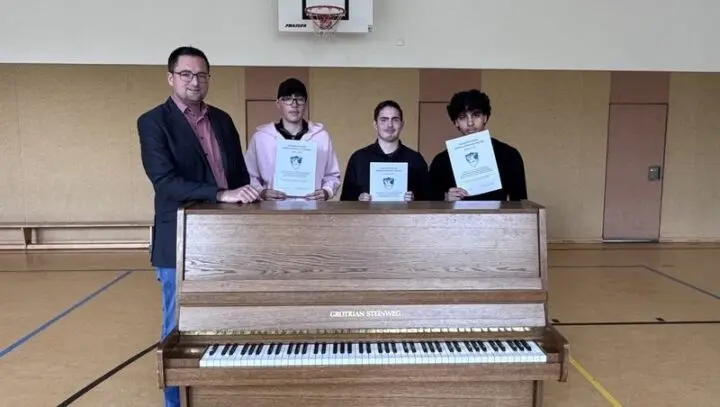 Drei Schüler mit Urkunden und ein Erwachsener stehen hinter einem Klavier in der Turnhalle