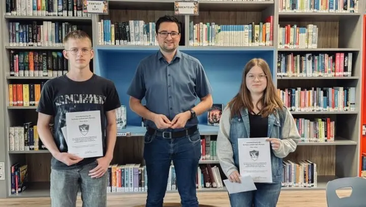 Schüler und Schülerin mit Urkunden, dazwischen ein Erwachsener vor Bücherregalen der Schülerbibliothek