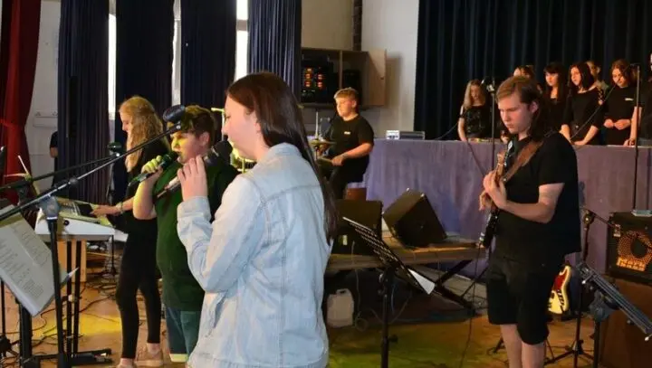 Schulband mit Sängerinnen und Sängern, Gitarrist und Schlagzeuger auf der Bühne, dahinter der Chor