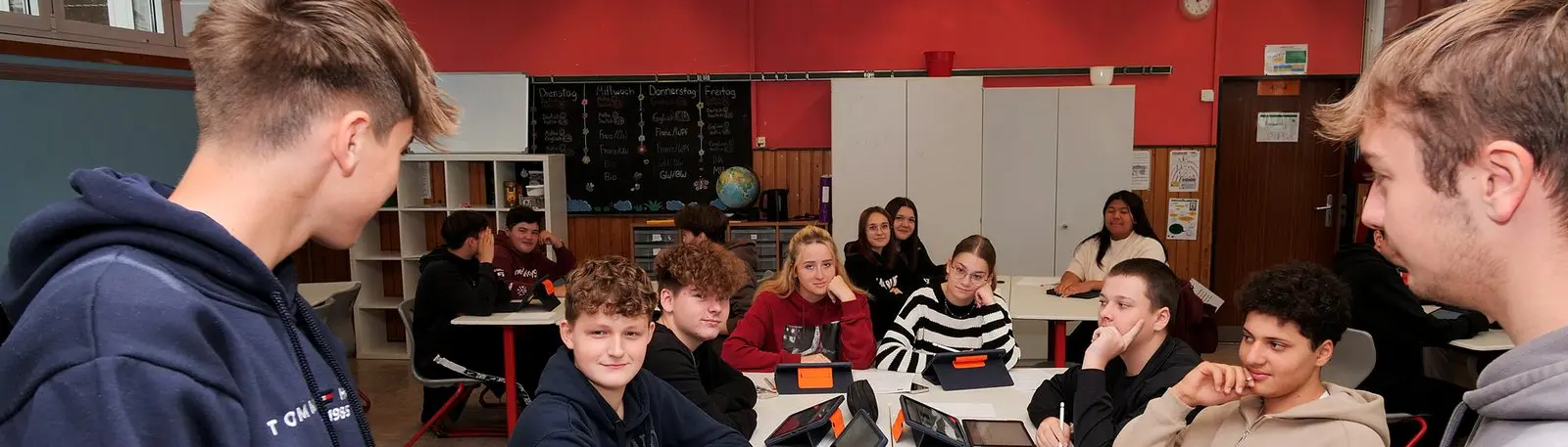 Zwei Jugendliche stehen vor einer Klasse, die an Gruppentischen mit Tablets sitzt