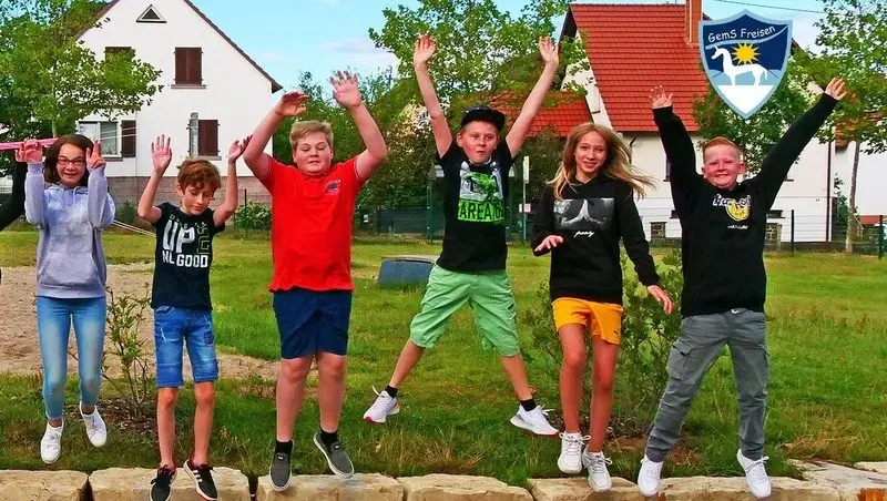 Sieben Jugendliche springen jubelnd mit erhobenen Armen von einer Steinmauer, oben rechts das Schullogo