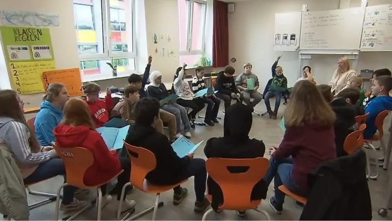 Schülerinnen und Schüler sitzen im Stuhlkreis mit Arbeitsblättern, einige melden sich, Standbild aus einem Fernsehbericht