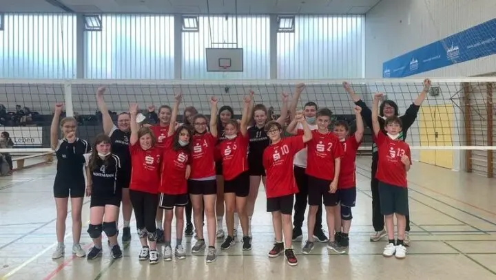 Jubelnde Mannschaft in roten Trikots mit erhobenen Fäusten vor dem Volleyballnetz in der Sporthalle