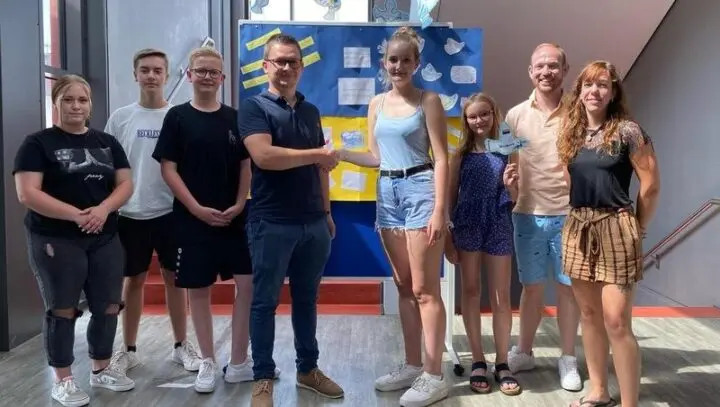 Gruppe vor blau-gelber Stellwand mit Papiertauben, ein Mann und eine Schülerin geben sich die Hand