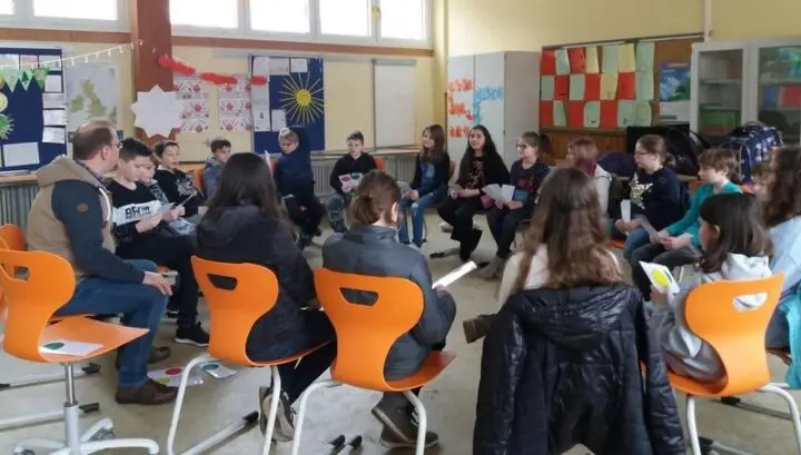 Schülerinnen und Schüler sitzen mit einem Erwachsenen im Stuhlkreis und halten Karten in den Händen