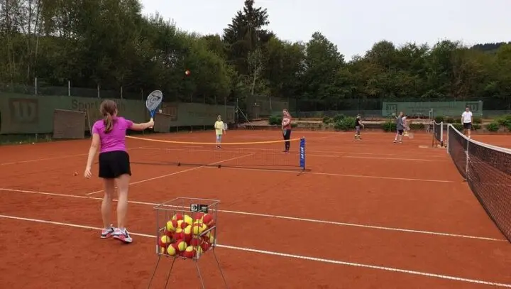 Schülerinnen und Schüler beim Tennisspielen auf einem Sandplatz, vorne ein Korb mit Bällen