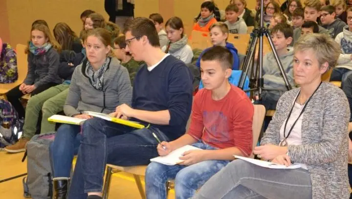 Vier Personen mit Bewertungsbögen in der ersten Reihe, dahinter zuhörende Schülerinnen und Schüler