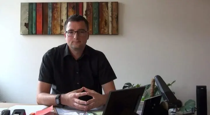 Mann mit Brille sitzt an einem Schreibtisch vor einem bunten Wandbild, Standbild aus einem Video