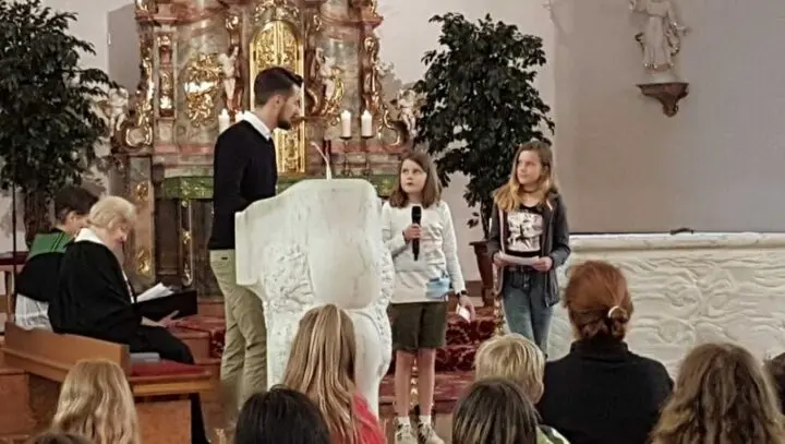 Zwei Mädchen am Ambo in der Kirche, eines spricht ins Mikrofon, daneben ein Erwachsener