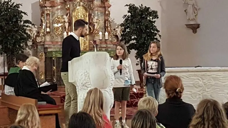 Zwei Mädchen am Ambo in der Kirche, eines spricht ins Mikrofon, daneben ein Erwachsener