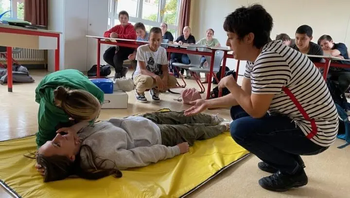 Jugendliche prüft bei einer liegenden Mitschülerin die Atmung, eine Ausbilderin erklärt, die Klasse schaut zu