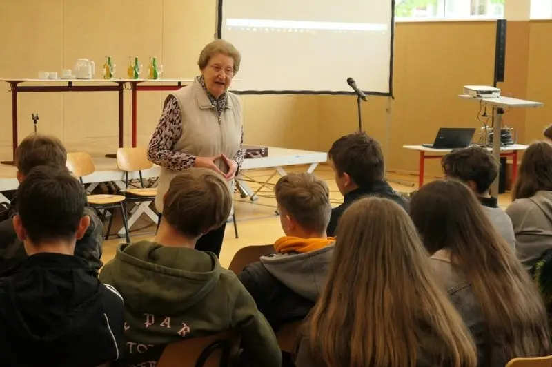 Eine Frau spricht im Saal zu sitzenden Schülerinnen und Schülern, dahinter eine Leinwand
