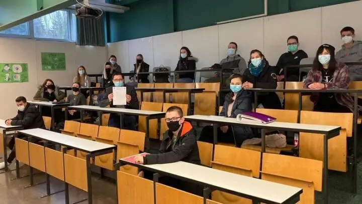 Schülerinnen und Schüler mit Masken sitzen mit Abstand im Hörsaal, einer hält sein Zeugnis hoch