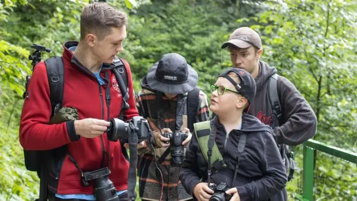 Jugendliche mit Kameras im Wald, einer zeigt einem Jungen etwas an seiner Kamera