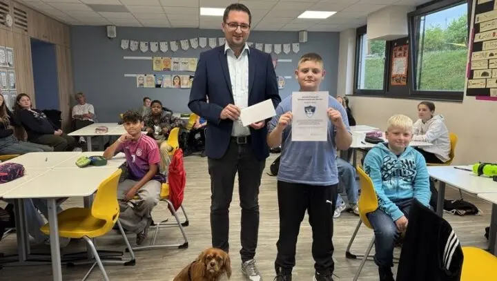 Schüler hält die Urkunde „Schülervorbild des Monats“ hoch, daneben ein Erwachsener mit Umschlag im Klassenraum