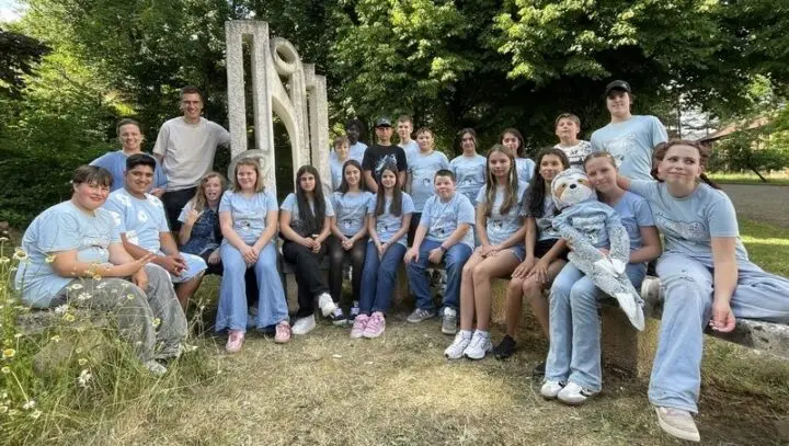 Gruppe von Schülerinnen und Schülern in hellblauen T-Shirts vor einer Steinskulptur, eine hält ein Plüschfaultier