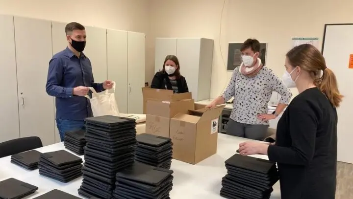 Vier Erwachsene mit Masken an einem Tisch mit gestapelten Tablets und Versandkartons