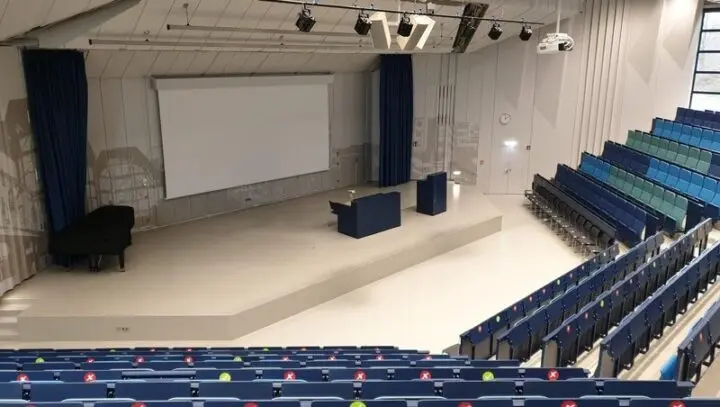 Leerer Hörsaal mit ansteigenden blauen Sitzreihen, Bühne, Rednerpult und Flügel